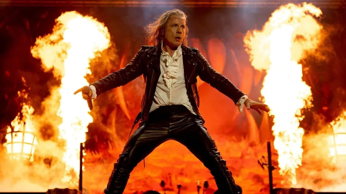 Bruce Dickinson performing live on stage with arms outstretched, dramatic flames erupting behind him during an Iron Maiden concert.