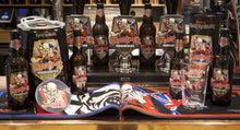 Load image into Gallery viewer, Display of Iron Maiden Trooper Premium British Beer merchandise, including bottles, pint glasses, a mini keg, pump clips, and coasters featuring Eddie the Trooper mascot, along with branded bar mats, crafted by Robinsons Brewery.
